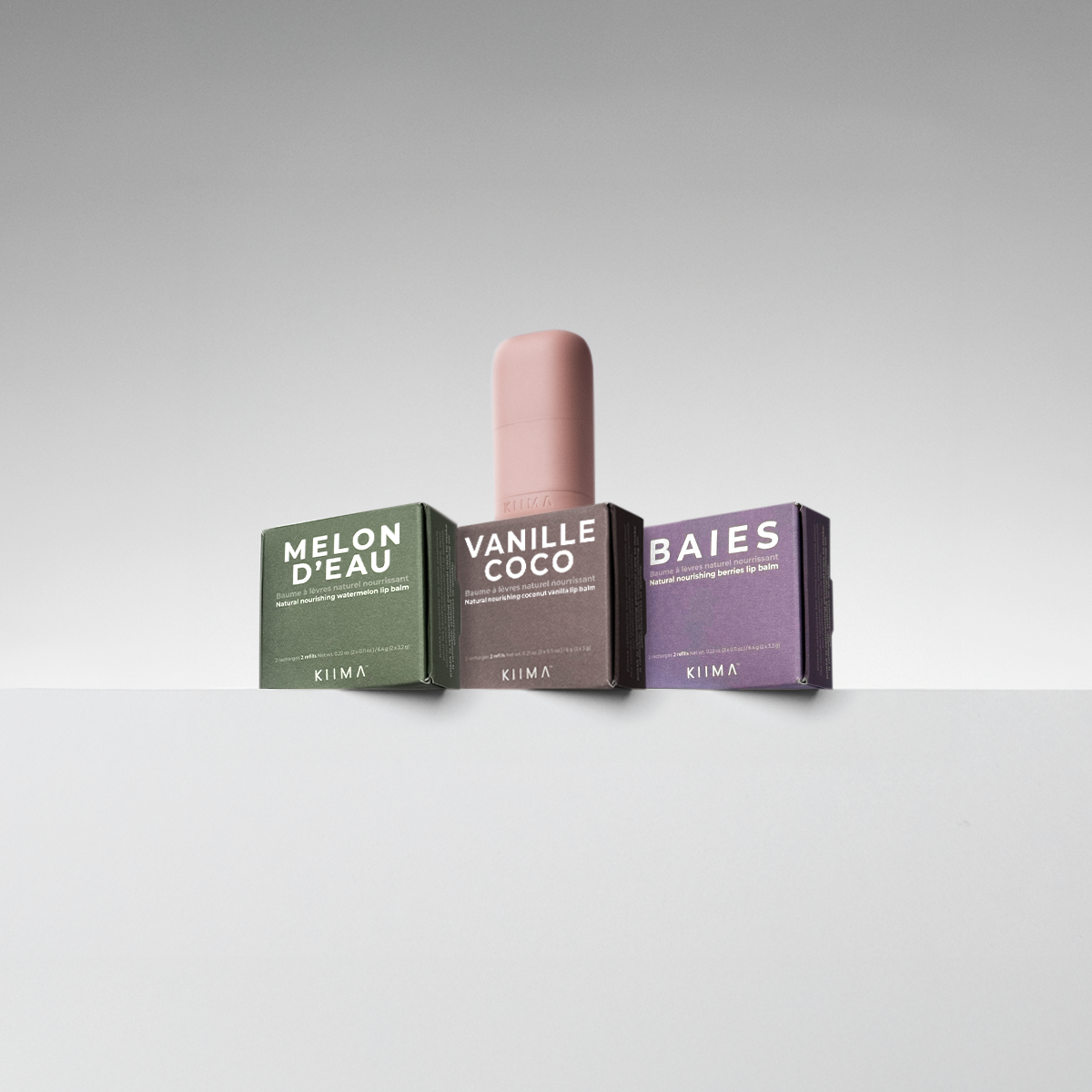 Three KIIMA product boxes labelled 'Melon d'Eau', 'Vanille Coco', and 'Baies' with pink refillable lip balm on a white surface against a gray background.