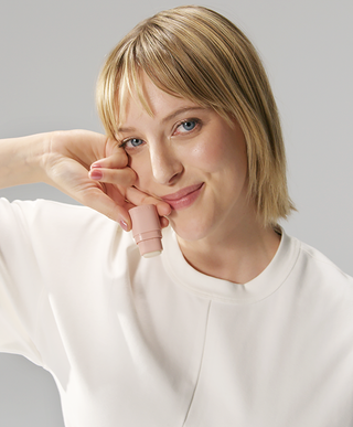 A blonde woman with blue eyes wearing white clothing, holding a pink KIIMA refillable lip balm upside down near her mouth against a minimalist grey background.