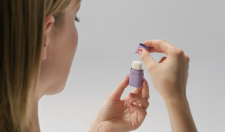 A woman demonstrates how to use a KIIMA refillable lip balm by removing the cap from a small purple applicator.