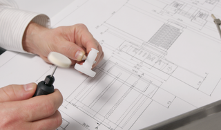 Hands using a precision engraving tool on a KIIMA refillable deodorant applicator prototype sketch.