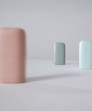 A collection of KIIMA refillable lip balms in green, blue, and pink, displayed with a shallow depth of field against a minimalist grey background to highlight the various color options.
